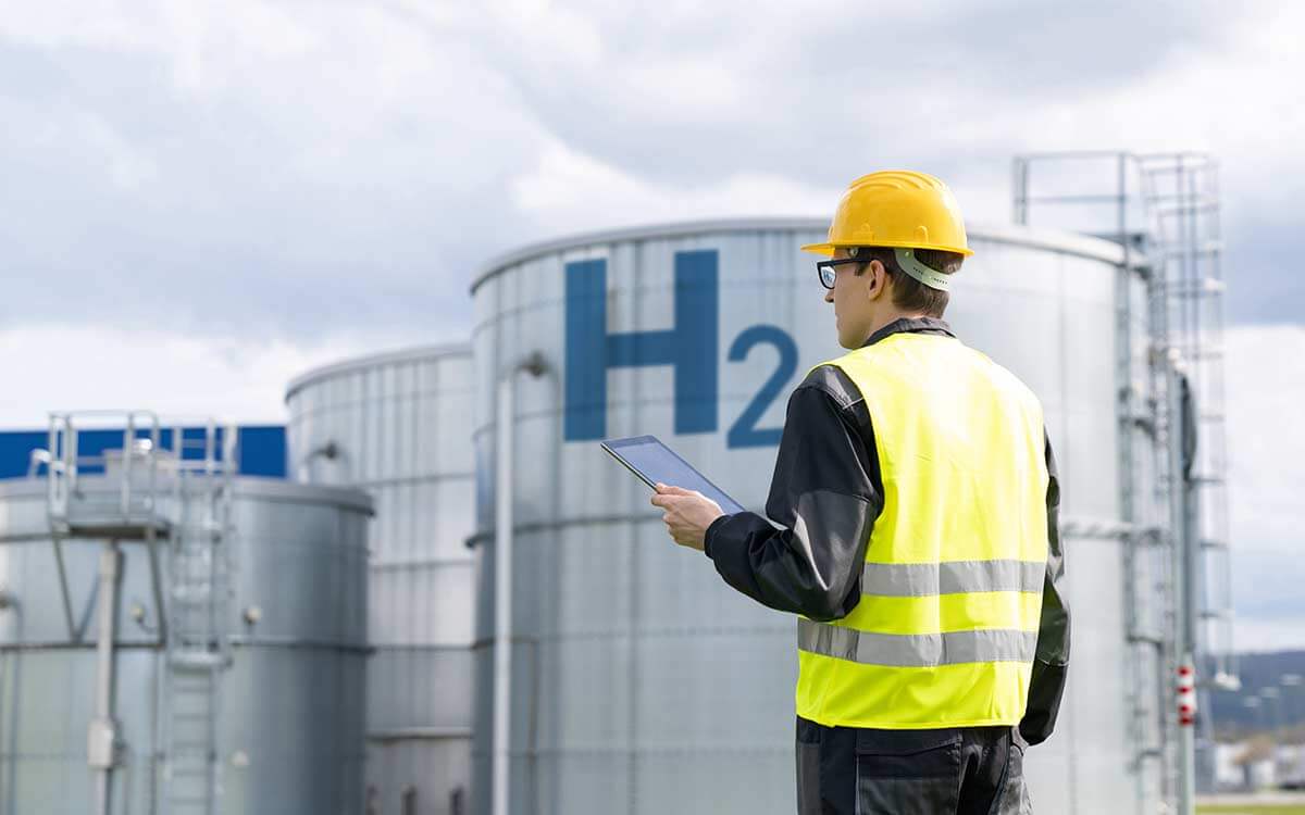 Ein Mann mit gelbem Arbeitshelm und Warnweste steht vor einem Wasserstoffspeicher