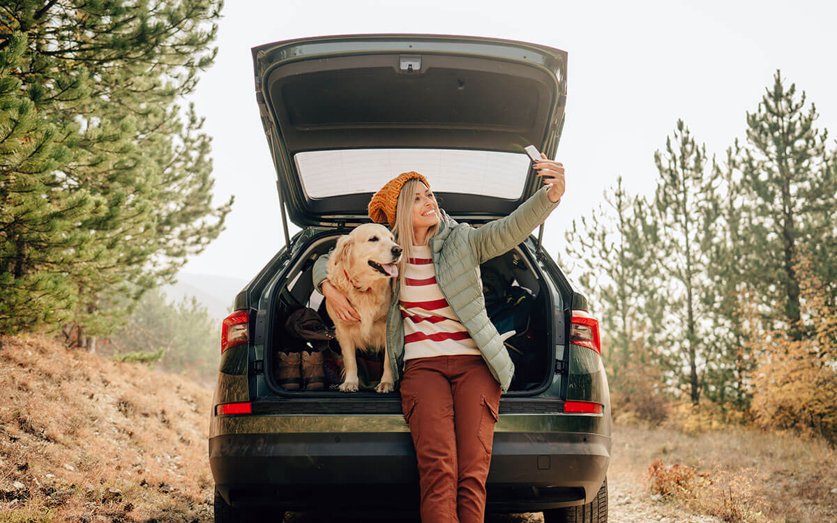 Eine Frau macht ein Selfie von sich und ihrem Hund im geöffneten Heck ihres Autos sitzend
